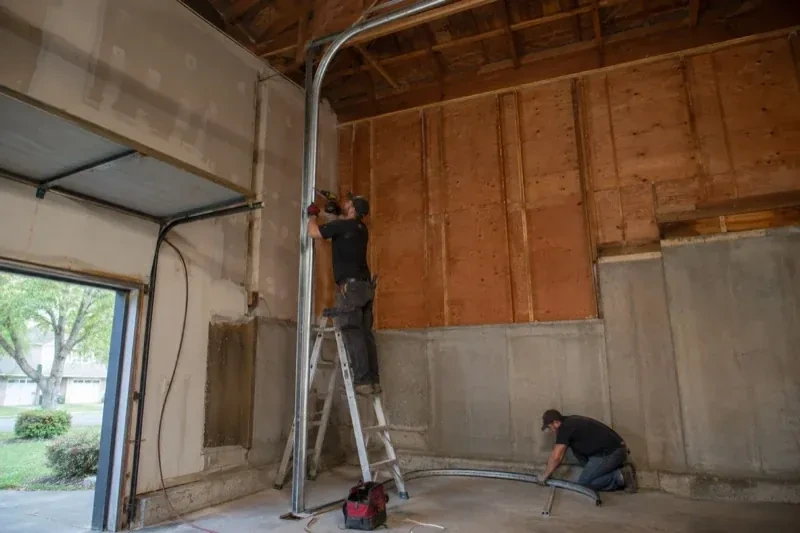 One person on a stepladder working beside a tall curved garage-door track while another kneels beside loose track sections in an unfinished garage. — High-Lift Garage Door Conversion, Springwater